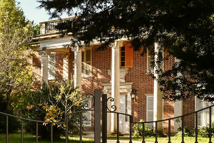 A colonnaded brick house in Uruguay, seen through iron gates and framed by mature trees